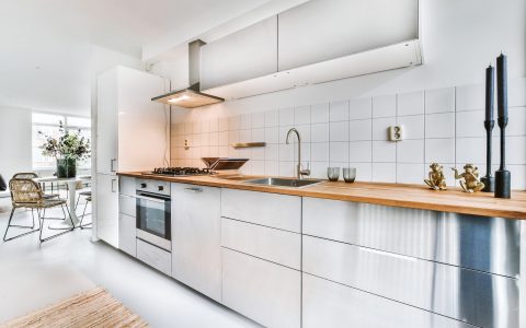 Modern home open plan kitchen with white tiles and cabinets and wooden counter equipped with stove and hood in contemporary apartment